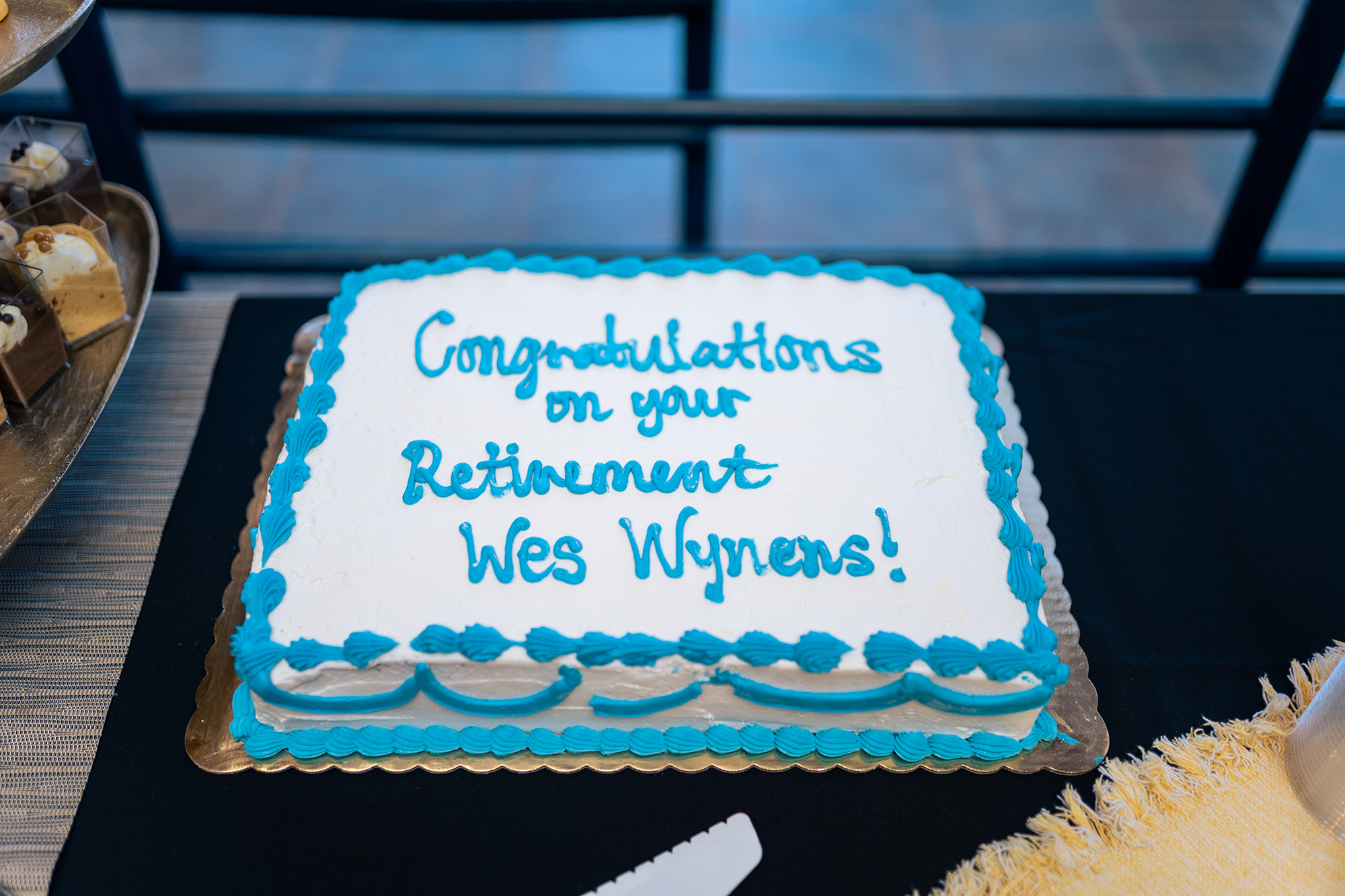 White cake with writing in blue frosting saying Congratulations on your retirement Wes Wynens