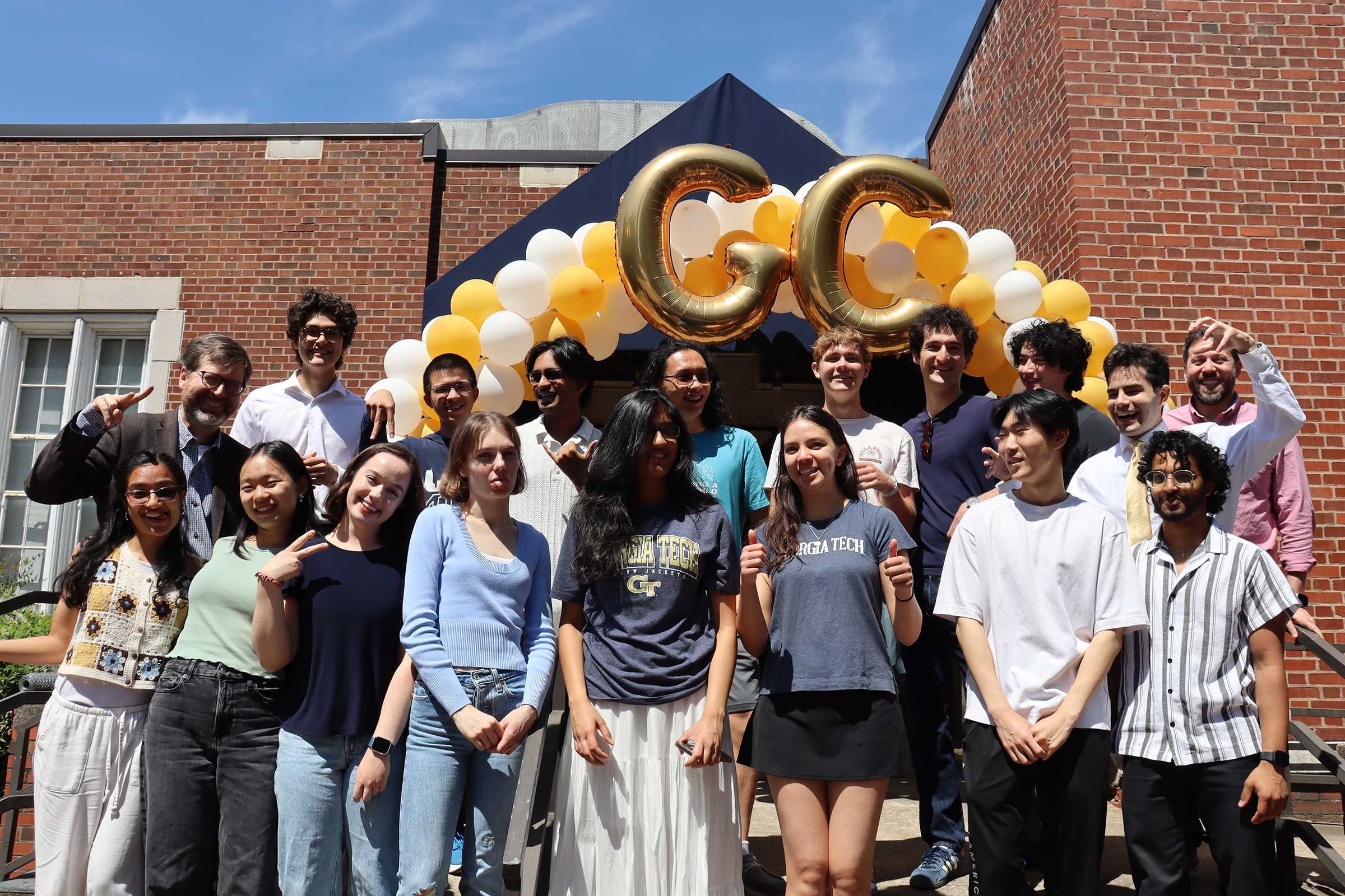 a large group of college students standing together under an arch of gold and white balloons