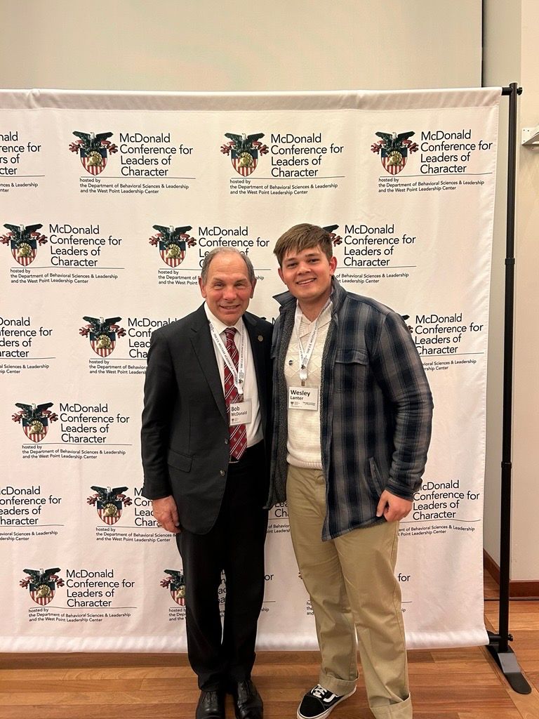 Image of Wesley Lanter (right) and MCLC Conference Founder, Bob McDonald (left) smiling in front of a banner that displays the conference name and West Point logo