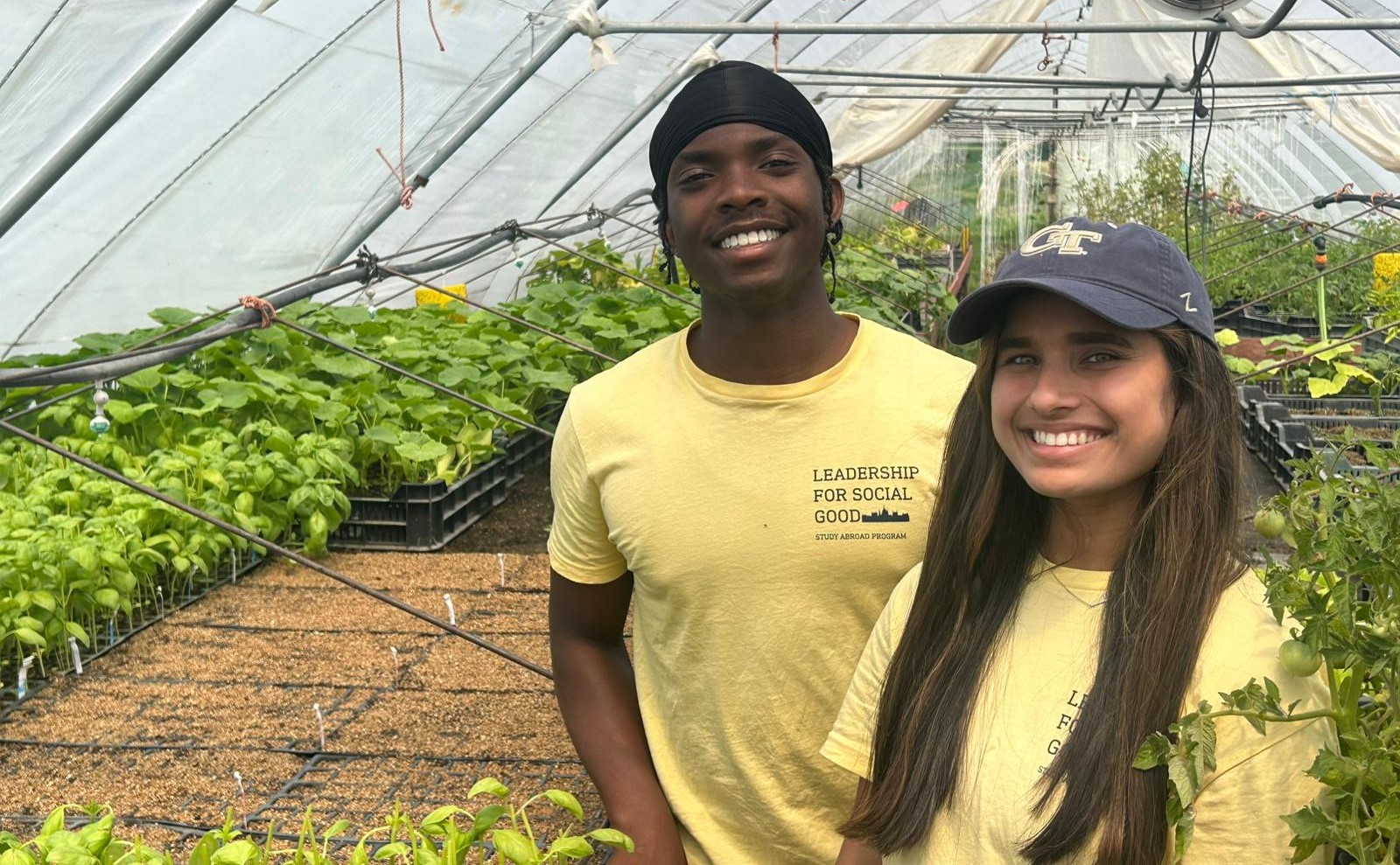 Two students in yellow Leadership for Social Good shirts stand inside a greenhouse, surrounded by rows of young plants.