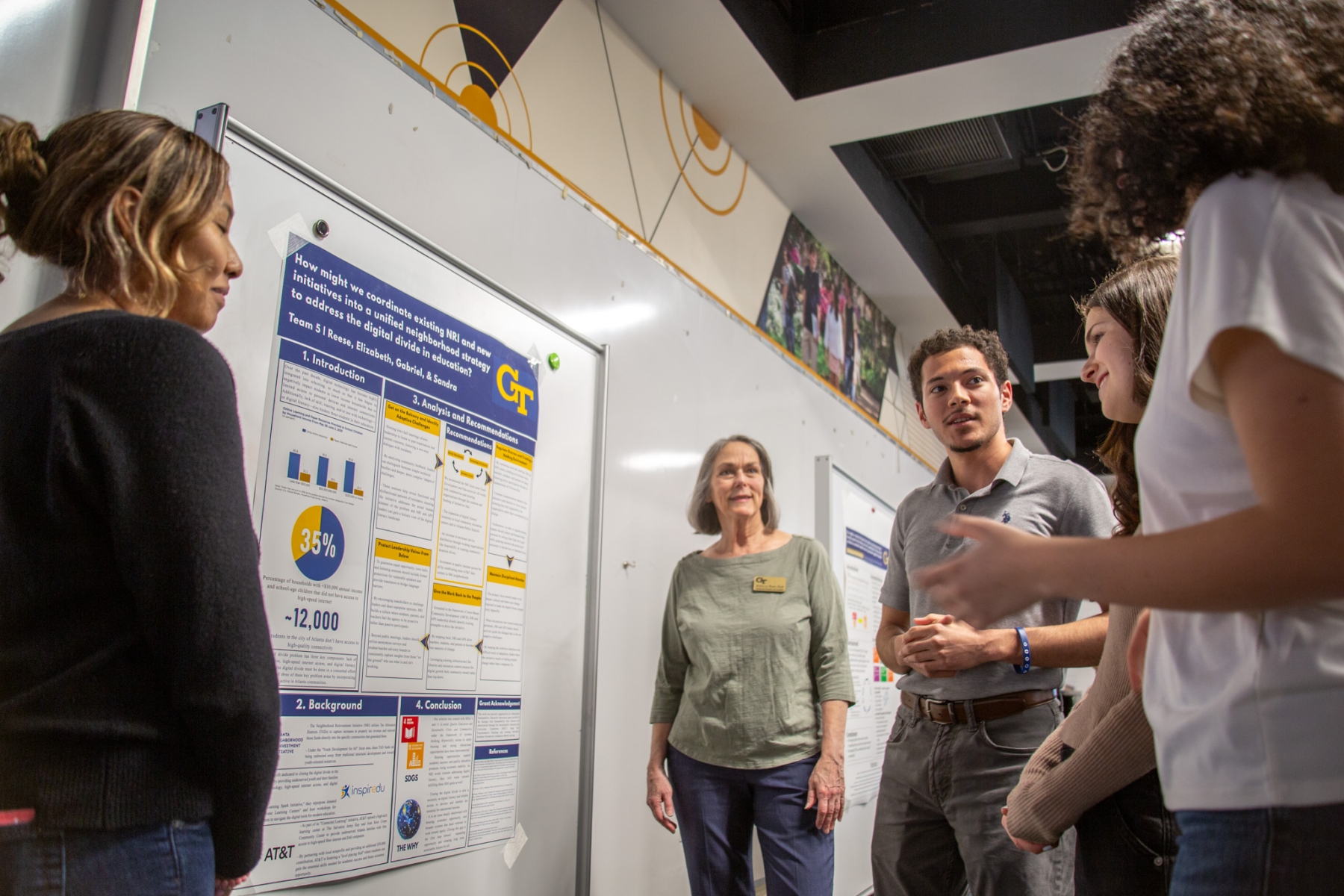 Students present and discuss a research poster on a whiteboard while a small group stands nearby and listens in a classroom setting..