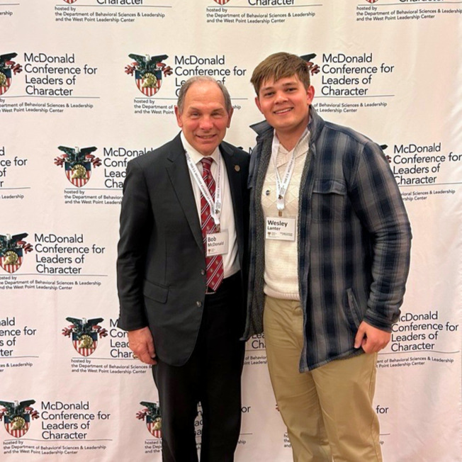 Wesley Lanter (right) and MCLC Conference Founder, Bob McDonald smiling in front of a banner that displays the conference name and West Point logo 