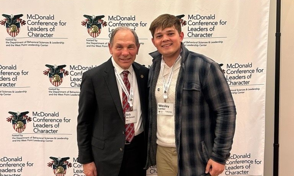Image of Wesley Lanter (right) and MCLC Conference Founder, Bob McDonald (left) smiling in front of a banner that displays the conference name and West Point logo