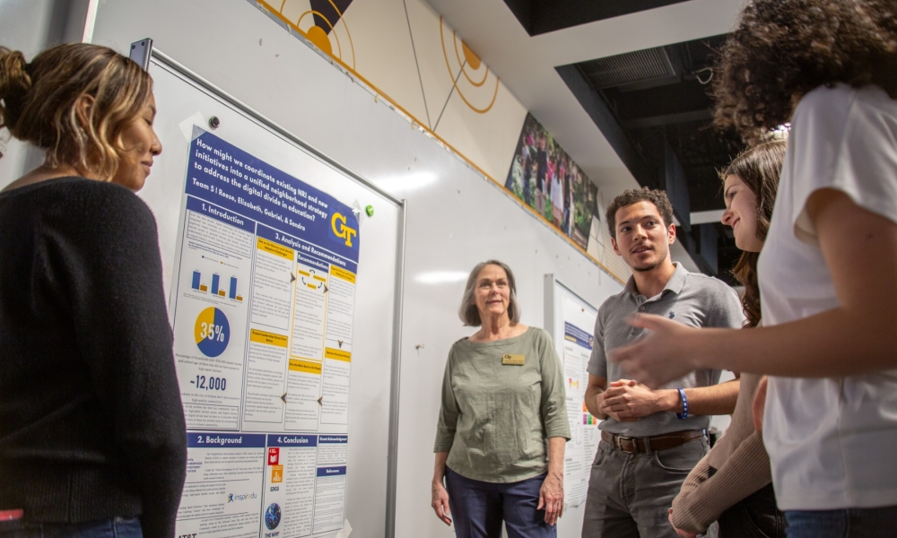 Students present and discuss a research poster on a whiteboard while a small group stands nearby and listens in a classroom setting..