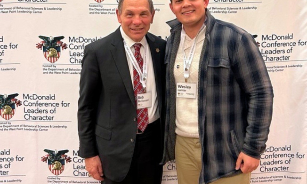 Wesley Lanter (right) and MCLC Conference Founder, Bob McDonald smiling in front of a banner that displays the conference name and West Point logo 