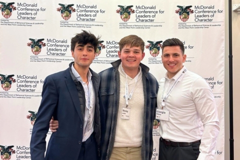 Wesley Lanter (center) and two other conference attendees smiling in front of a banner that displays the conference name and West Point logo