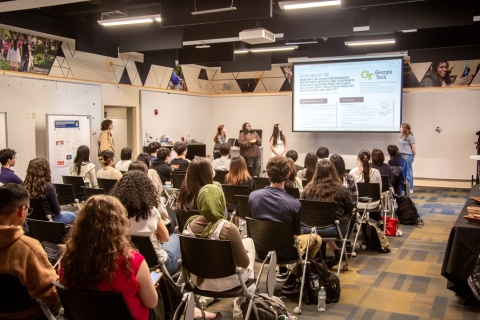 Students sit in a classroom while a few students speak at the front of the room in front of a screen displaying a presentation slide.